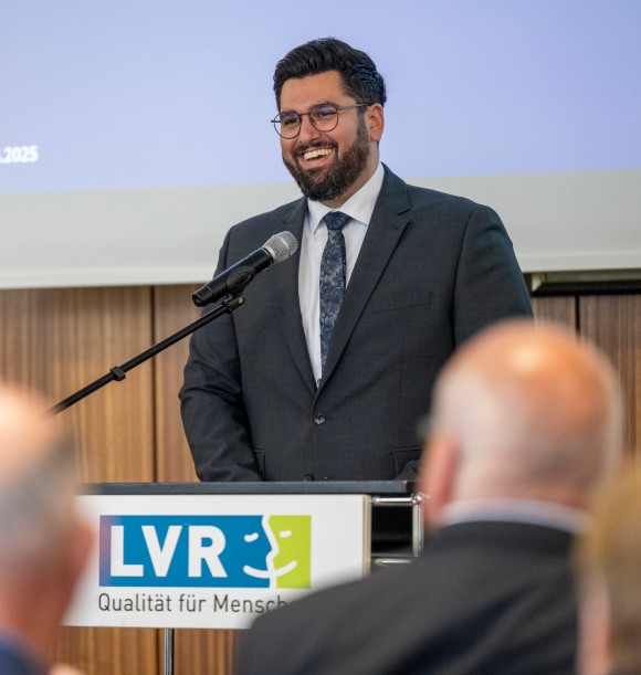 Man sieht einen Mann im Anzug, der einen schwarzen Blazer trägt. Er steht an einem Redepult, an dem vorne das LVR-Logo zu sehen ist.