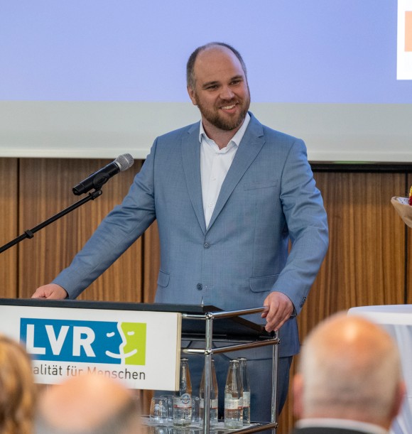 Man sieht einen Mann mit blauem Anzug, der hinter einem Redepult steht. An dem Redepult ist vorne das LVR-Log zu sehen.