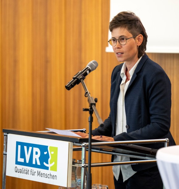 Man sieht eine Frau, die an einem Pult steht und eine Rede hält. An dem Redepult ist vorne das LVR-Log zu sehen.