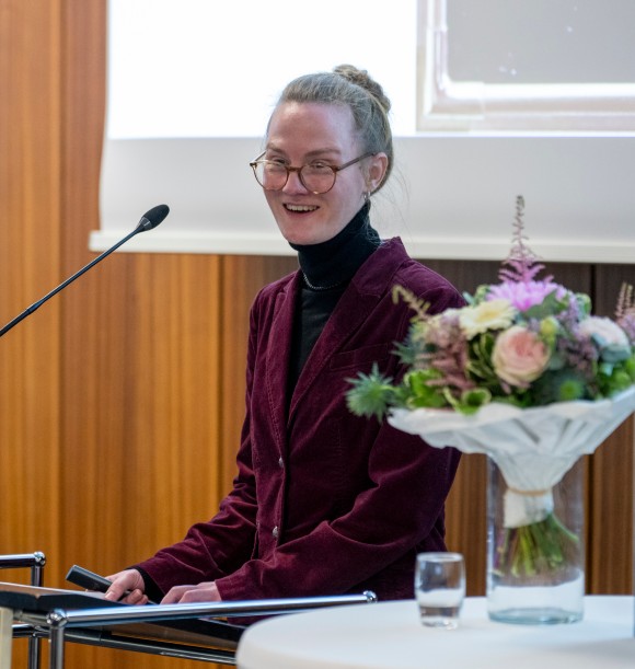 Eine Frau in einem weinroten Blazer an einem Rednerpult.