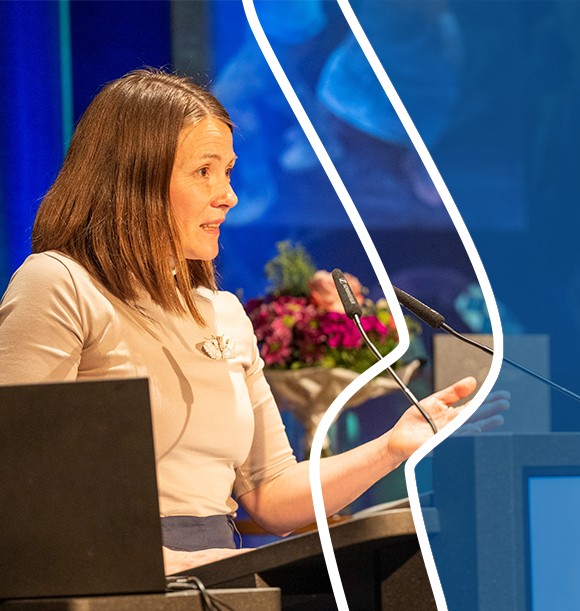 Man sieht eine brünette Frau aus dem Profil, die an einem Redepult steht und in ein Mikrofon spricht. 