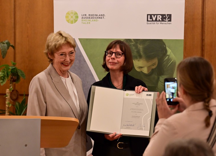 Zwei Frauen stehend, eine Frau vor einem Pult, die andere Frau hält eine Urkunde.