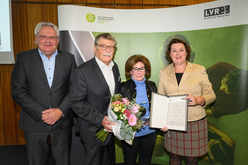 Man sieht zwei Männer in Anzügen und zwei Frauen in formeller Kleider vor dem grünen Aufsteller des Rheinlandtalers des LVR. Einer der Männer hält einen Blumenstrauß in den Händen. Eine der Frauen hält die Trophäe des Rheinlandtalers und die andere die Urkunde des Rheinlandtalers in den Händen.