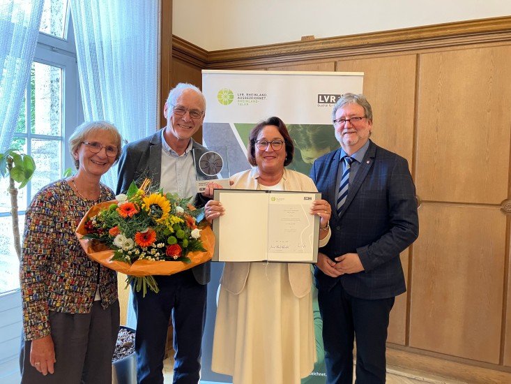 Eine Gruppe von vier festlich gekleideten, lächelnden Menschen. In der Mitte steht ein Mann, der eine Trophäe hält, eine Frau die einen Blumenstrauß träägt und eine Urkunde wird gezeigt.