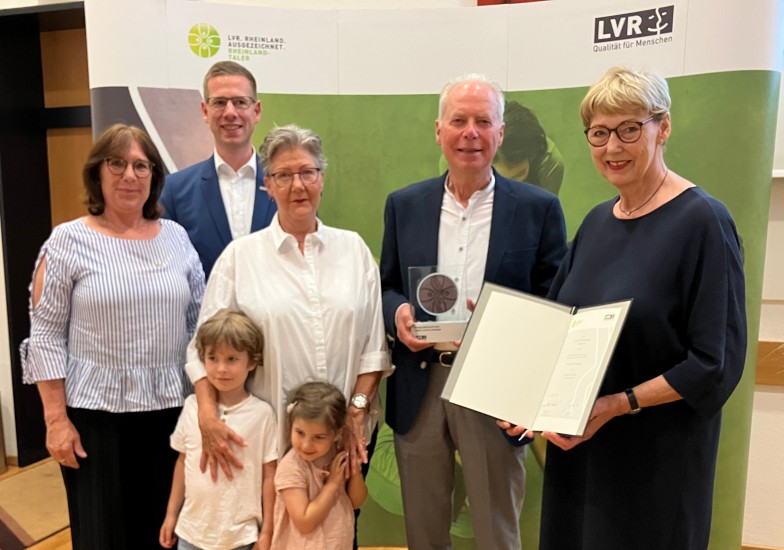 Ein Gruppe festlich gekleideter Menschen, die lächelnd vor einem Banner mit dem LVR-Logo stehen. Die Frau ganz links hält eine Urkunde in der Hand. Der Mann links neben ihr hält eine Trophäe.