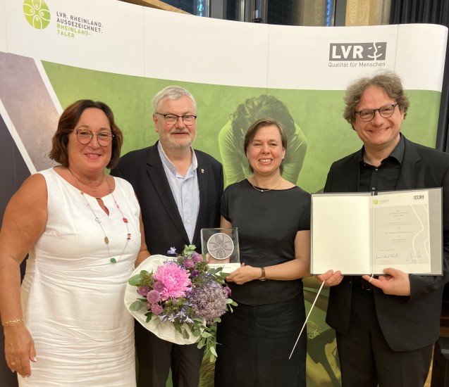 Ein Gruppe festlich gekleideter Menschen, die lächelnd vor einem Banner mit dem LVR-Logo stehen. Der Mann ganz rechts hält eine Urkunde in der Hand. Die Frau links neben ihm hält eine Trophäe.