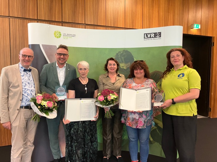 Eine Gruppe festlich gekleideter Menschen in einem Saal. Der Mann und eine Frau halten einen Blumenstrauss, zwei Frauen haltene eine Urkunde und ein Mann und eine Frau halten eine Trophäe in der Hand.