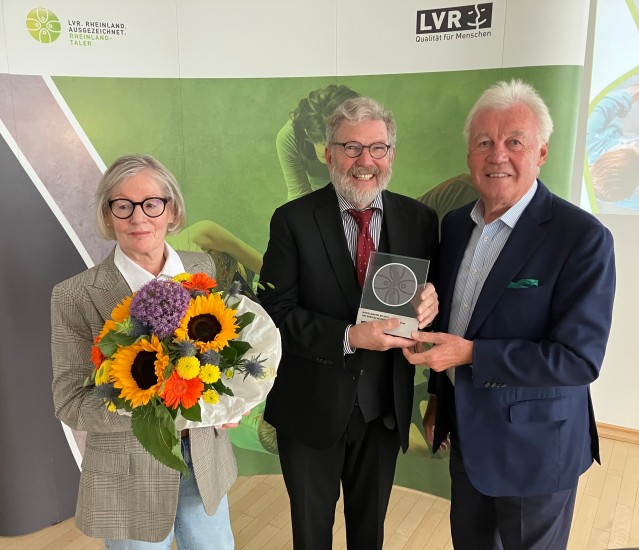Ein Gruppe von drei festlich gekleideten Menschen, die Frau links hält einen Blumenstrauß und der Mann in der Mitte hält eine Trophäe.