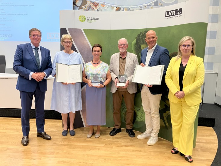 Ein Gruppe festlich gekleideter Menschen, die in die Kamera lächeln. Zwei Personen halten jeweils eine Urkunde und die beiden in der Mitte halten Trophäen in den Händen.
