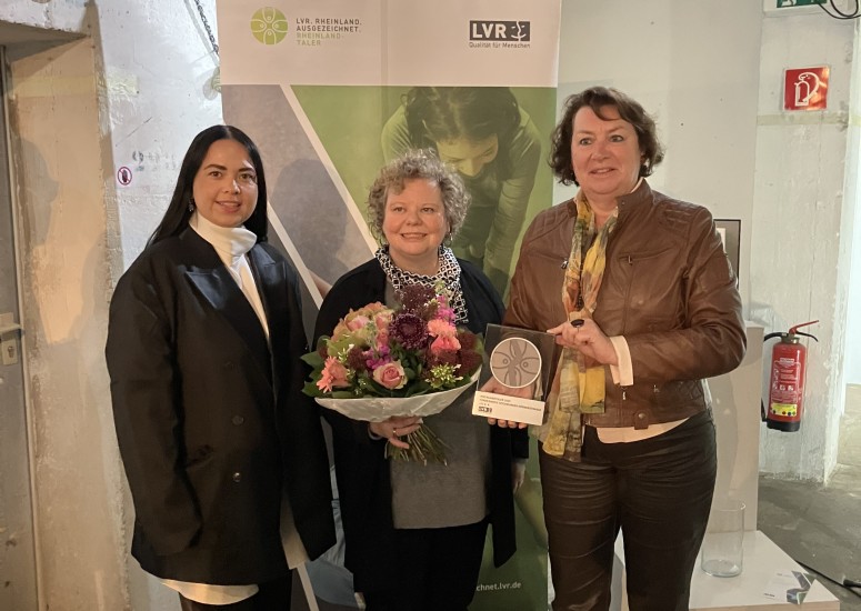 Drei Frauen stehen vor einer Leinwand mit der Aufschrift 