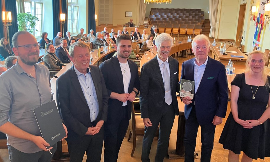Eine Gruppe festlich gekleideter Menschen in einem Saal. Der zweite Mann von rechts hält eine Trophäe in der Hand.