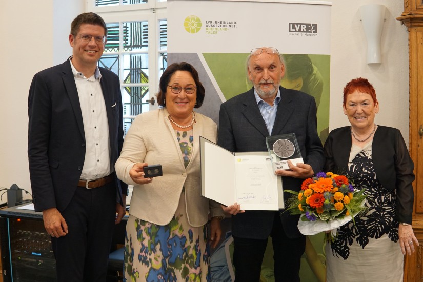 Eine Gruppe von vier festlich gekleideten, lächelnden Menschen. In der Mitte steht ein Mann, der eine Trophäe hält, eine Frau die einen Blumenstrauß trägt und eine Urkunde wird gezeigt.