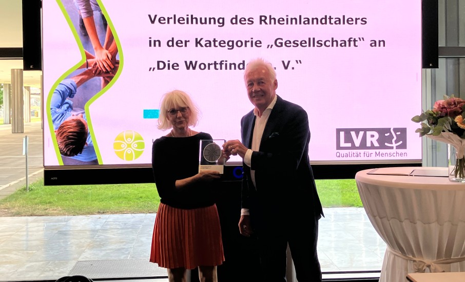 Eine festlich gekleidete Frau und ein Mann stehen vor einer Leinwand. Auf der Leinwand steht