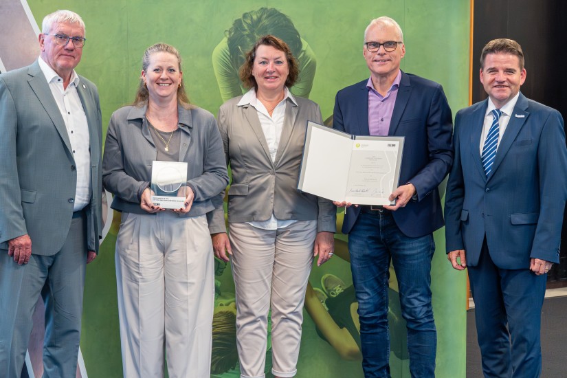 Eine Gruppe von fünf festlich gekleideter Personen vor einer Leinwand. Eine Frau (die zweite Person von links) hält eine Trophäe in der Hand. Eine (zweite Person von rechts) hält eine Urkunde hoch.