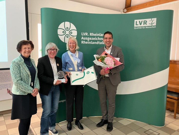 Drei Frauen und ein Mann stehen vor einem grünen Hintergrund mit dem Logo und dem Schriftzug des Rheinlandtalers. Die zweite Frau von links hält eine Trophäe in der Hand.