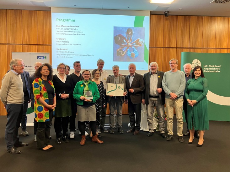Eine Gruppe Menschen, die vor einer Leinwand stehen. Eine Frau in der Mitte hält eine Trophäe in der Hand. Ein Mann deneben eine Urkunde.
