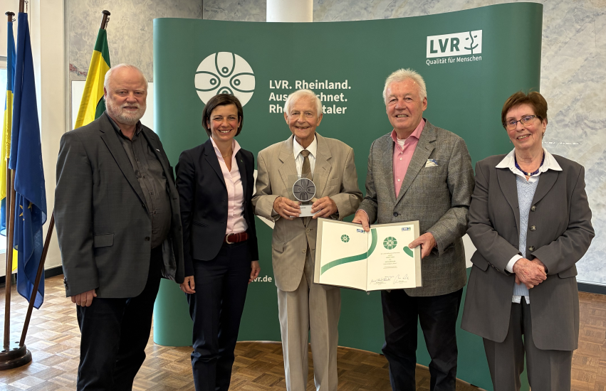 Eine Gruppe Menschen steht vor einem grünen Hintergrund mit dem Logo des Rheinlandtalers. Der Mann in der Mitte hält eine Trophäe in der Hand.