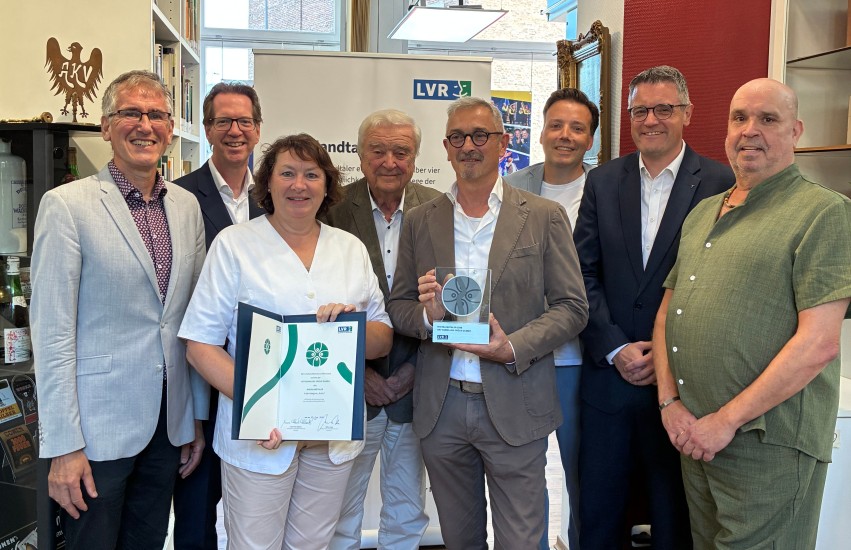 Ein Gruppenfoto. Eine Frau vorne hält eine Urkunde in der Hand. Ein Mann daneben eine Trophäe.