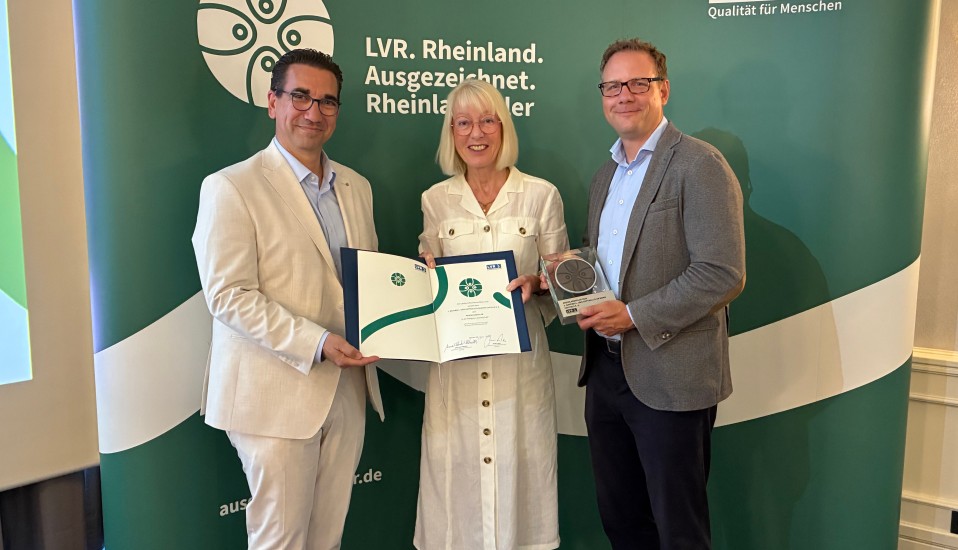 Zwei Männer und eine Frau vor einer grünen Leinwand. Die Frau in der Mitte hält eine Urkunde. Der Mann rechts daneben eine Trophäe.