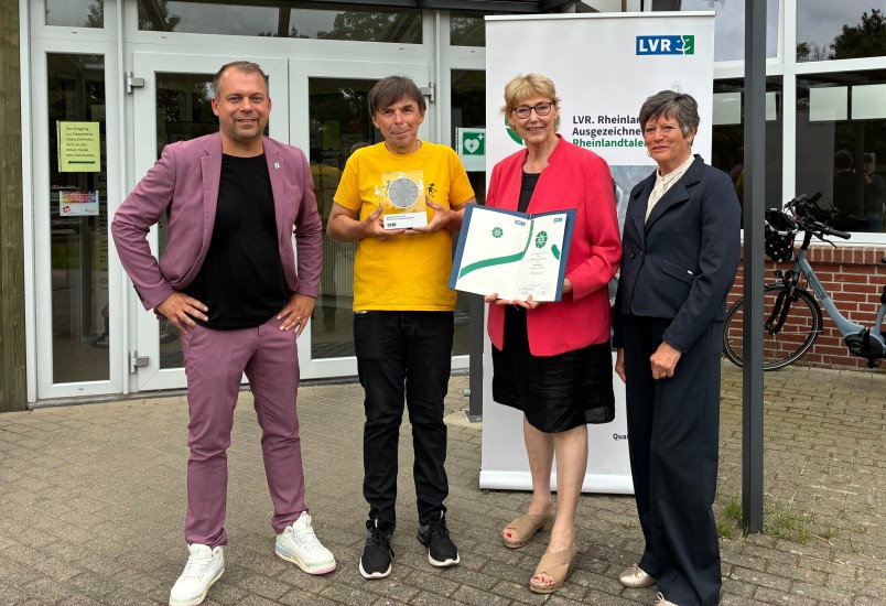 Drei Männer und eine Frau stehen vor einem Vereinsheim. Der zweite Mann von links hält eine Trophäe in der Hand.