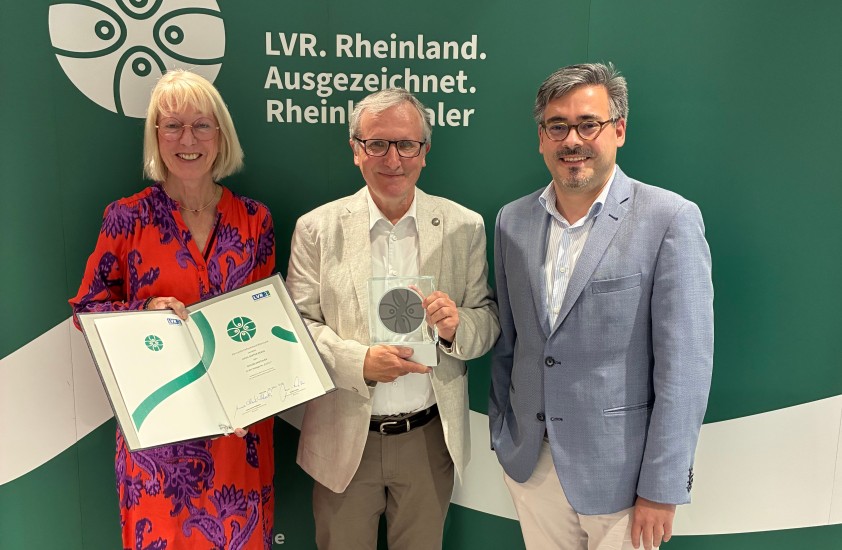 Eine Frau und drei Männer. Die Frau hält eine Urkunde in der Hand. Der Mann in der Mitte hält eine Trophäe.