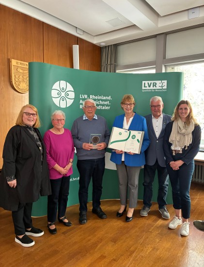 Drei Frauen und drei Männer stehend vor einer Wand; eine Frau hält Urkunde, ein Mann hält Trophäe